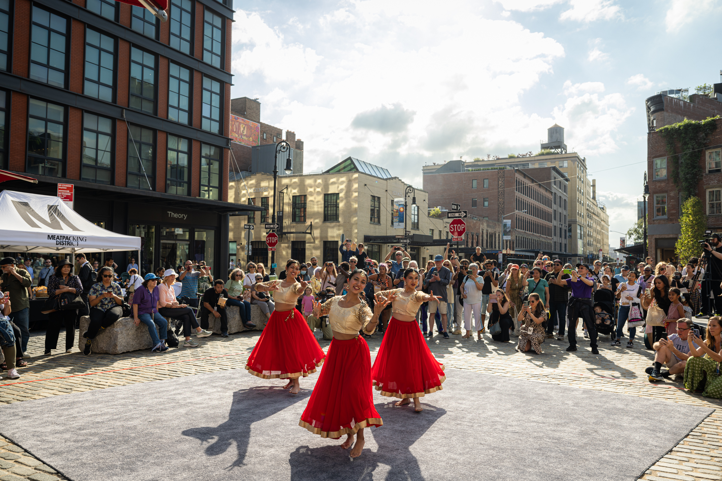 Three dancers in cream blouses and flowing red skirts perform barefoot on a gray outdoor carpet in the sunny Meatpacking District, surrounded by a large crowd watching and taking photos.