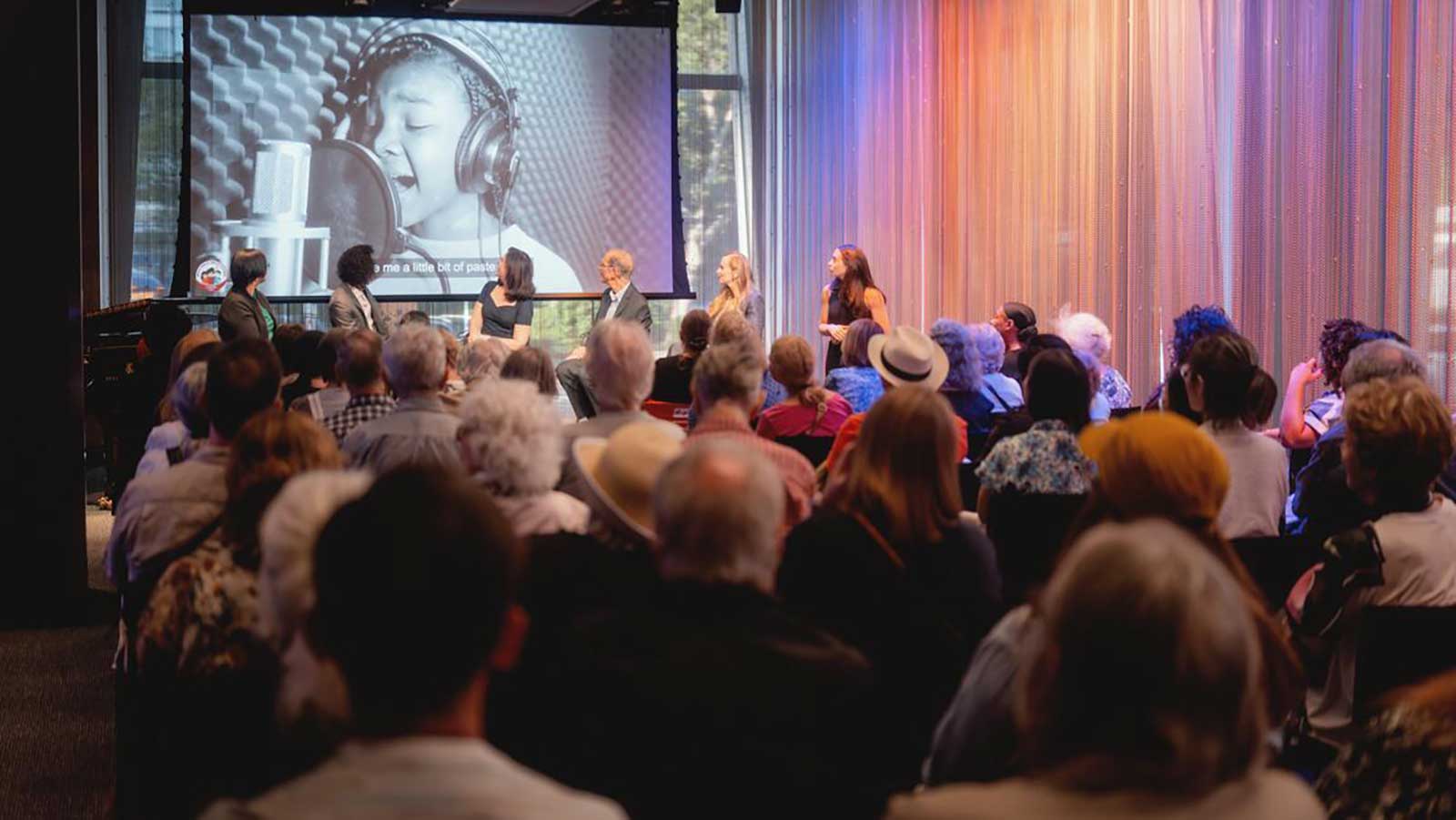 An audience looking towards a stage with six panelist turned around looking at a screen with a young women singing into a microphone