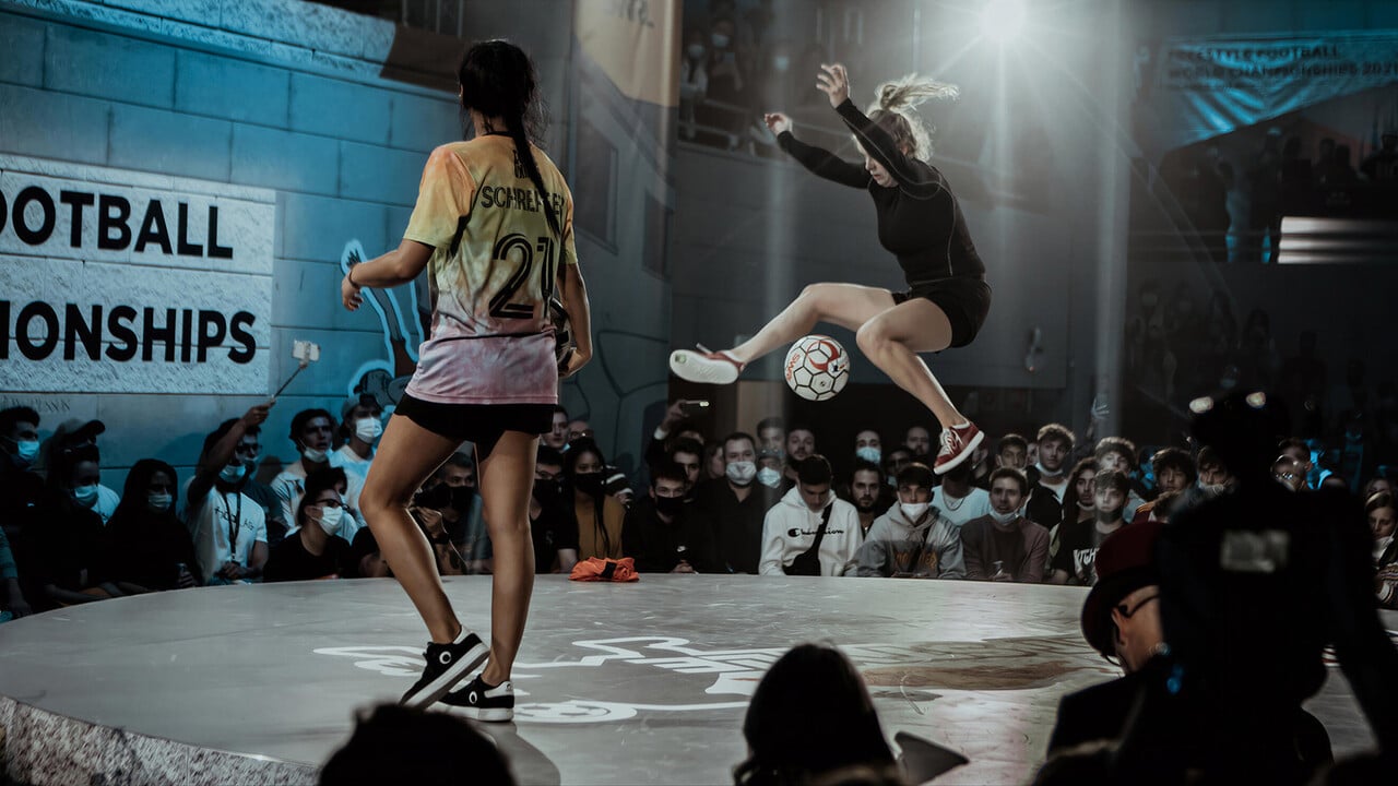 Two female soccer players doing freestyle soccer tricks on a stage with an audience.