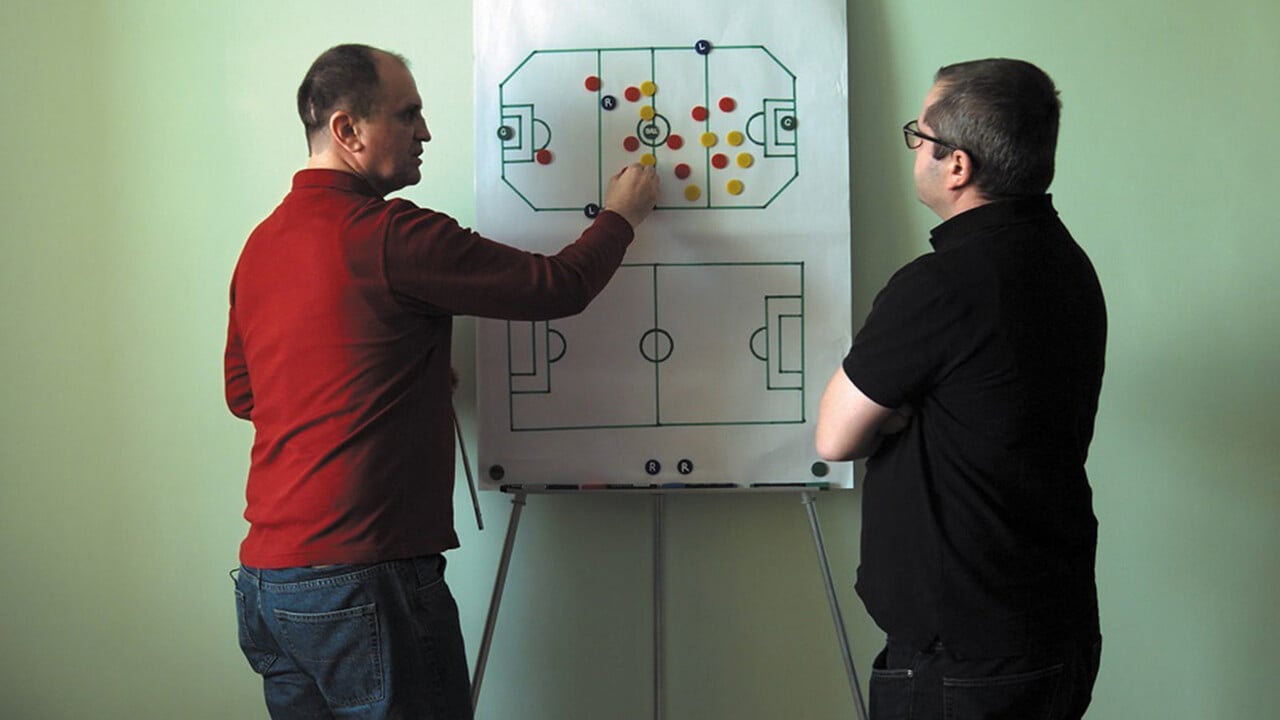 Two men standing infront of a play drawing, pointing at and moving pieces.